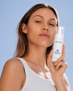 A woman in a white tank top holds a white amerin ESSENTIAL | Enzymatic Foaming Cleanser bottle against a clear blue sky background.