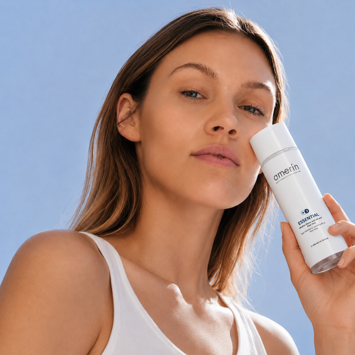A woman with a clear complexion holds a white AMERIN ESSENTIAL | Micellar Water with B5 and Aloe Vera bottle against a bright blue sky background.