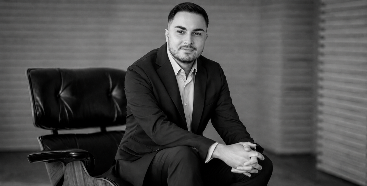 A black and white portrait of a Alexandru Albu, founder of amerin, in a dark suit and light dress shirt, seated in a black leather office chair against a modern slatted wall background.